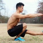 Homme en squat latéral, bras tendus, dans un parc, ciel gris, arbres sans feuilles