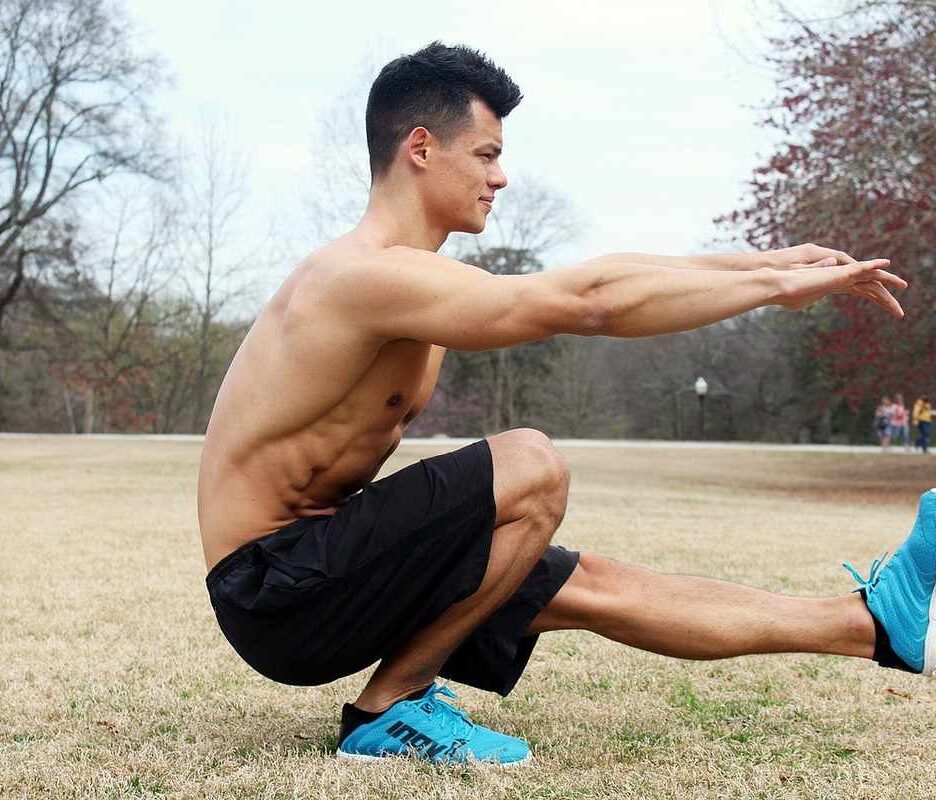 Homme en squat latéral, bras tendus, dans un parc, ciel gris, arbres sans feuilles