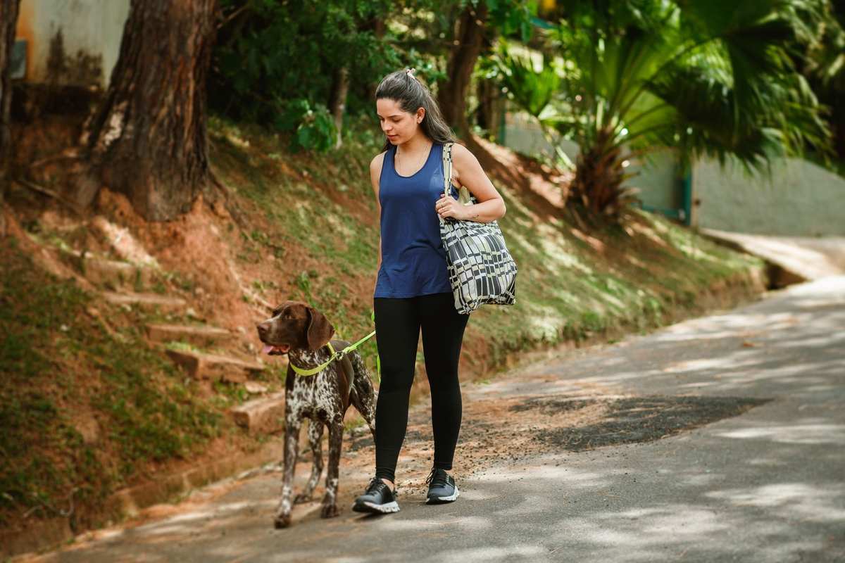 Femme marchant avec un chien sur un trottoir, sac à l'épaule, végétation dense en arrière-plan Femme marchant avec un chien sur un trottoir, sac à l'épaule, végétation dense en arrière-plan